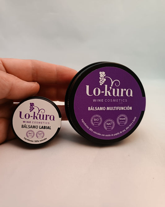 mano sujetando dos latas negras, una más pequeña con etiqueta beige y otra más grande con etiqueta morada en la que se lee lo-kura y bálsamo labial y multifunción sobre fondo neutro