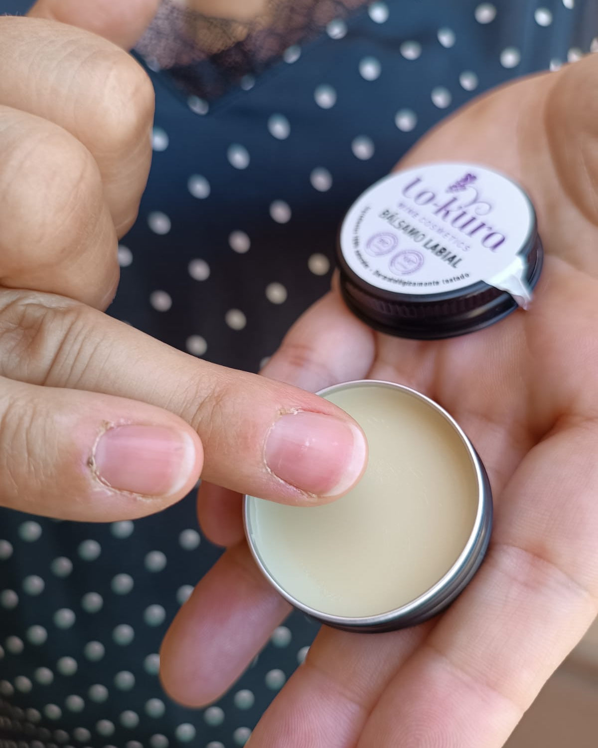 dedo sobre lata circular de metal negra con bálsamo labial dentro y tapa con etiqueta beige detrás.