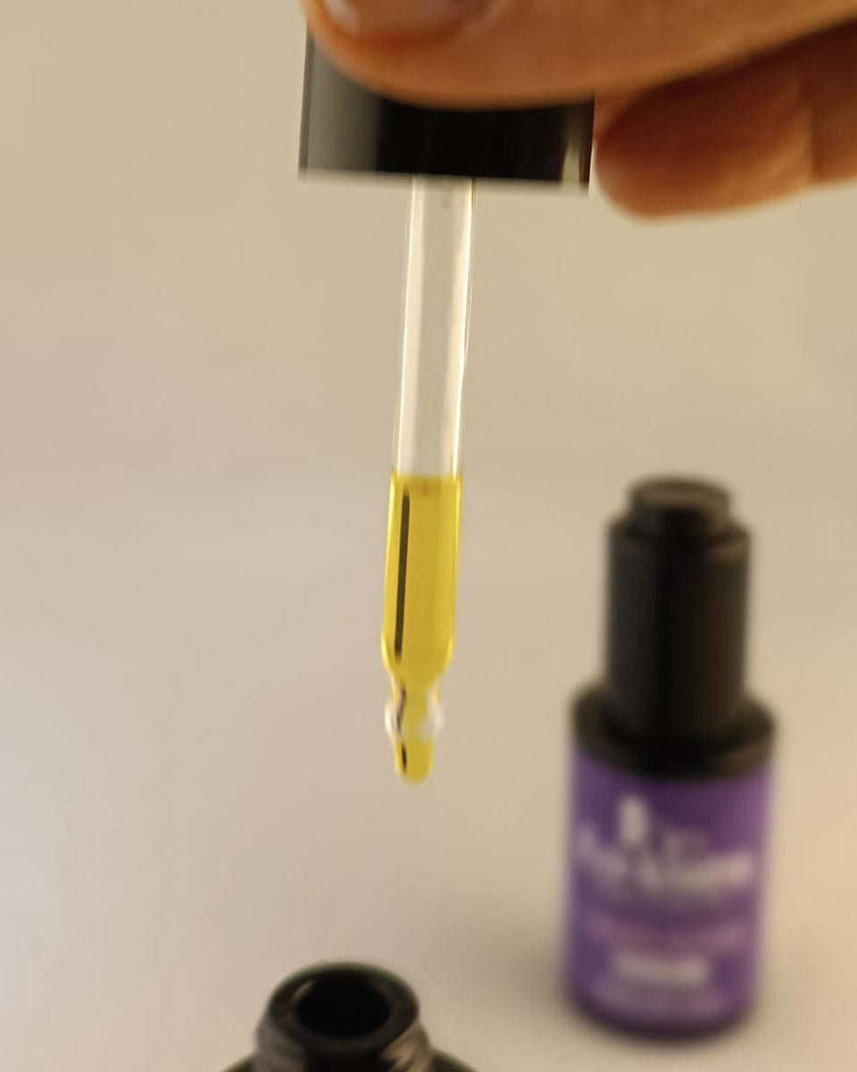 pipeta de cristal con líquido amarillo tipo aceite y una gota saliendo sobre boca de cristal negro y con fondo difuminado de frasco negro de cristal con etiqueta morada
