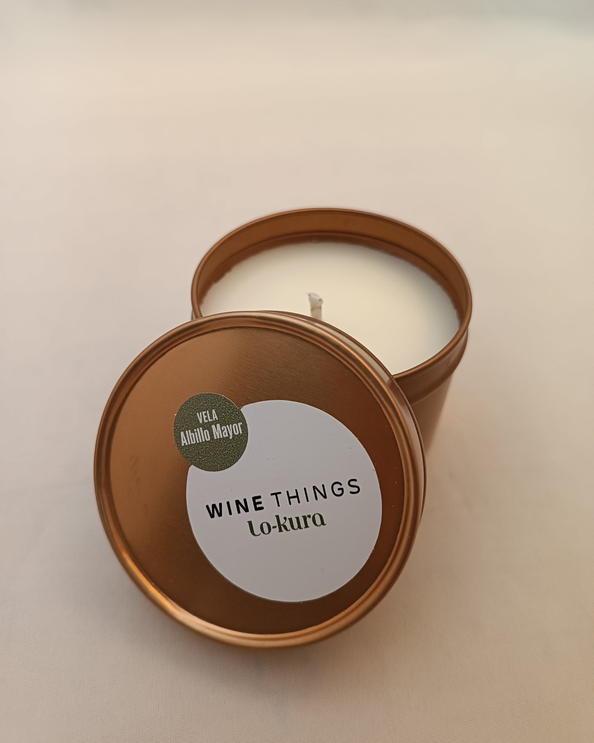 Vela en lata dorada de recipiente cilíndrigo con tapa apoyada en primer plano con pegatina donde se lee wine things lo-kura vela albillo mayor. fonodo neutro.