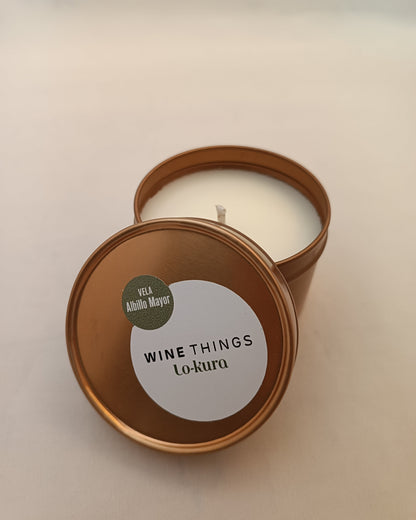 Vela en lata dorada de recipiente cilíndrigo con tapa apoyada en primer plano con pegatina donde se lee wine things lo-kura vela albillo mayor. fonodo neutro.